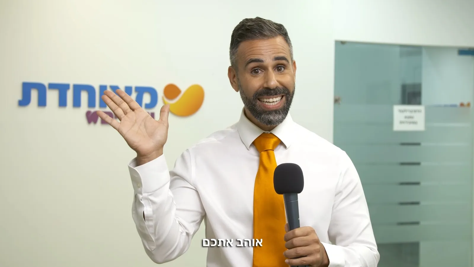 גבר עם זקן מחזיק מיקרופון ומחייך מול לוגו "מאוחדת", כתובית "אוהב אתכם" בתחתית הפריים