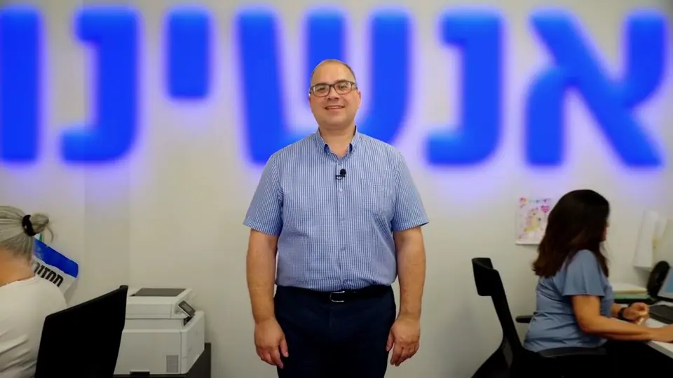 גבר עם מיקרופון דש מחייך במשרד פתוח, מאחוריו כיתוב כחול «אנשינו» ועובדים בעמדות