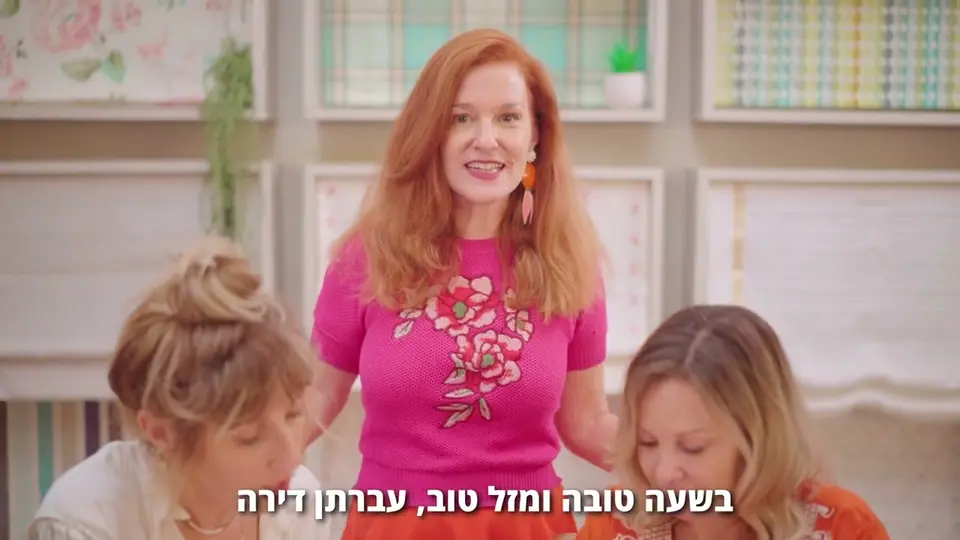 אישה ג׳ינג׳ית בסוודר ורוד מדברת בין שתי נשים, בסלון מעוצב עם דוגמאות בדים וטפטים