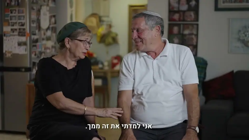 זוג מבוגר יושב יחד בסלון הבית, ידה של האישה על זרועו, ברקע תמונות משפחה על הקיר