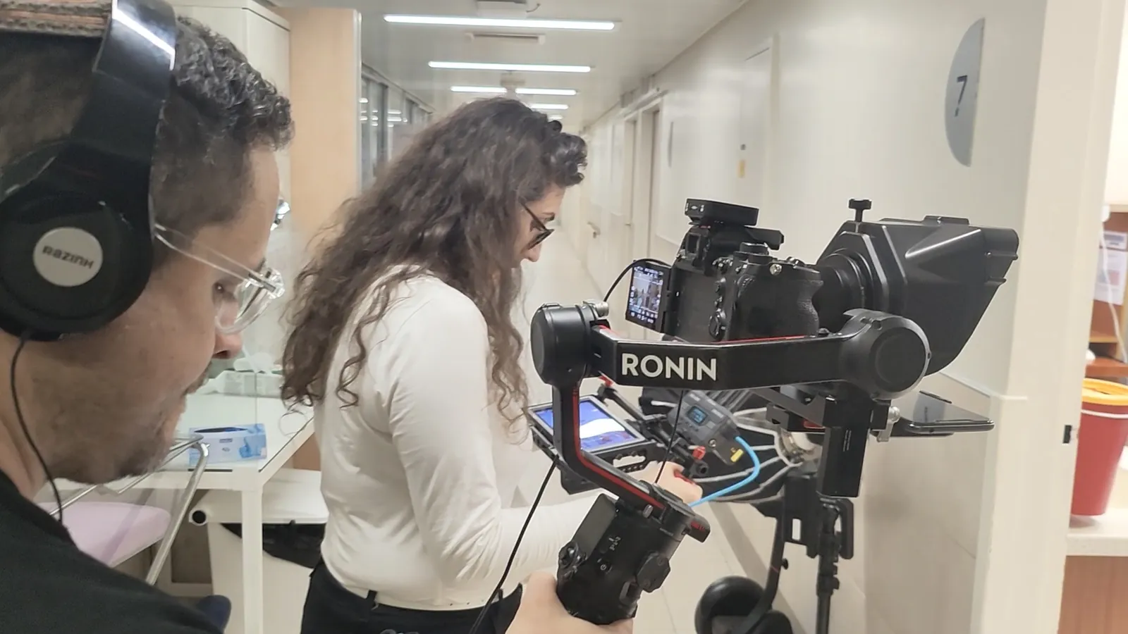 צלם עם אוזניות ומפיקה מכוונים מצלמה על גימבל RONIN עם תאורה ניידת במסדרון מחלקה בבית חולים